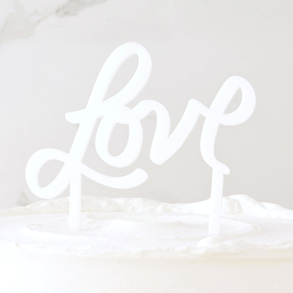 White acrylic cake topper that reads "love" in a hand drawn script on white frosted cake