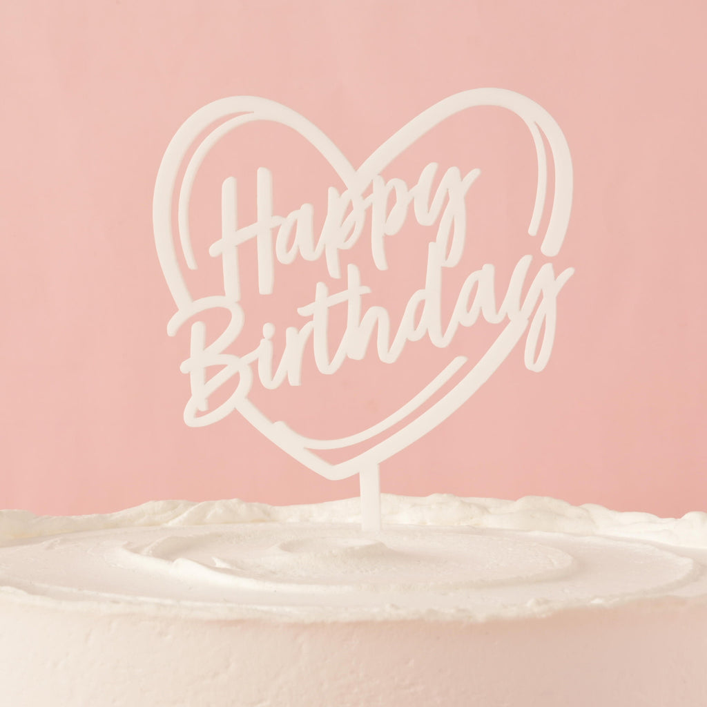 A white cake topper that says "Happy Birthday" inside a heart outline, placed on top of a frosted white cake. The background is a solid pink color.