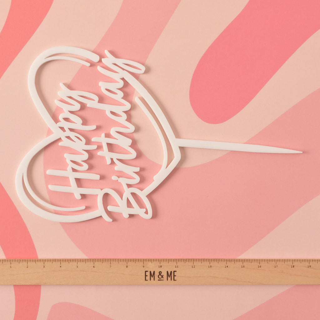 A close-up of a white cake topper that says "Happy Birthday" inside a heart outline. A ruler with the "EM & ME" logo is placed below the topper to show its size. The background is a pink surface with a wavy design.