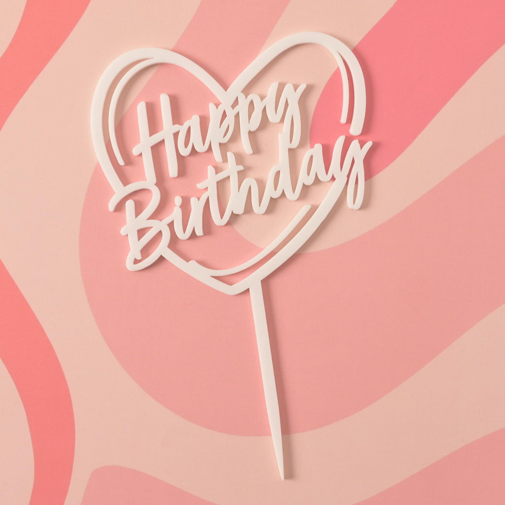 A close-up of a white cake topper that says "Happy Birthday" inside a heart outline. The topper is on a pink background with a wavy design.
