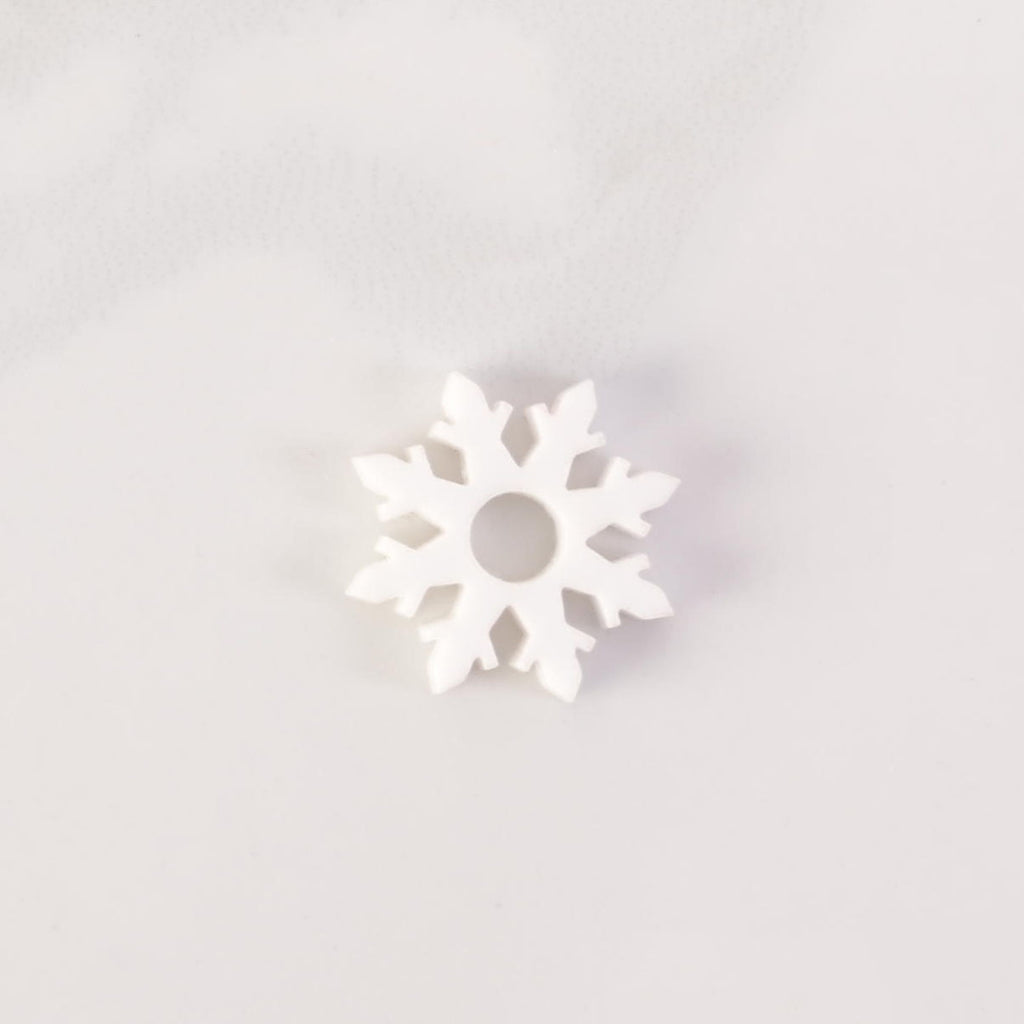 A close-up shot of a White snowflake-shaped charm with glitter accents and a hole in the middle. The charm is displayed on a white marble surface.