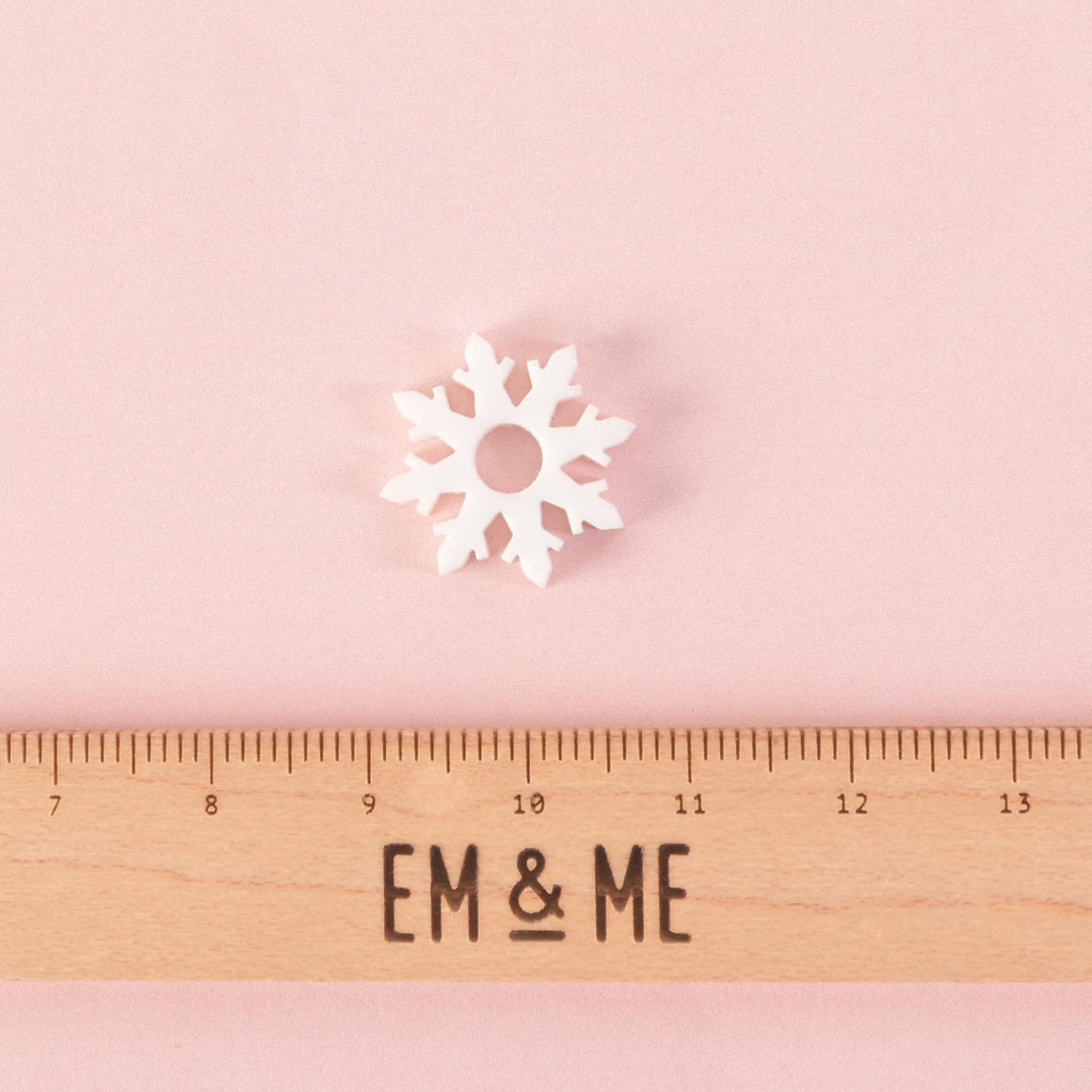 A single white snowflake-shaped charm or pin with a hole in the middle. A wooden ruler with the "EM & ME" logo is placed below the charm to show its size. The item is displayed on a solid pink background.
