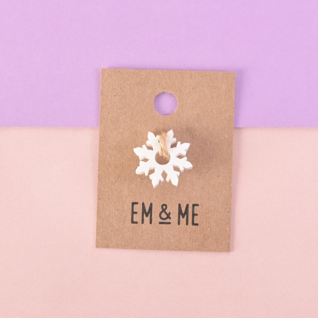 A white snowflake-shaped charm mini attached to a brown paper card that reads "EM & ME." The item is displayed on a background that is half solid purple and half solid pink.