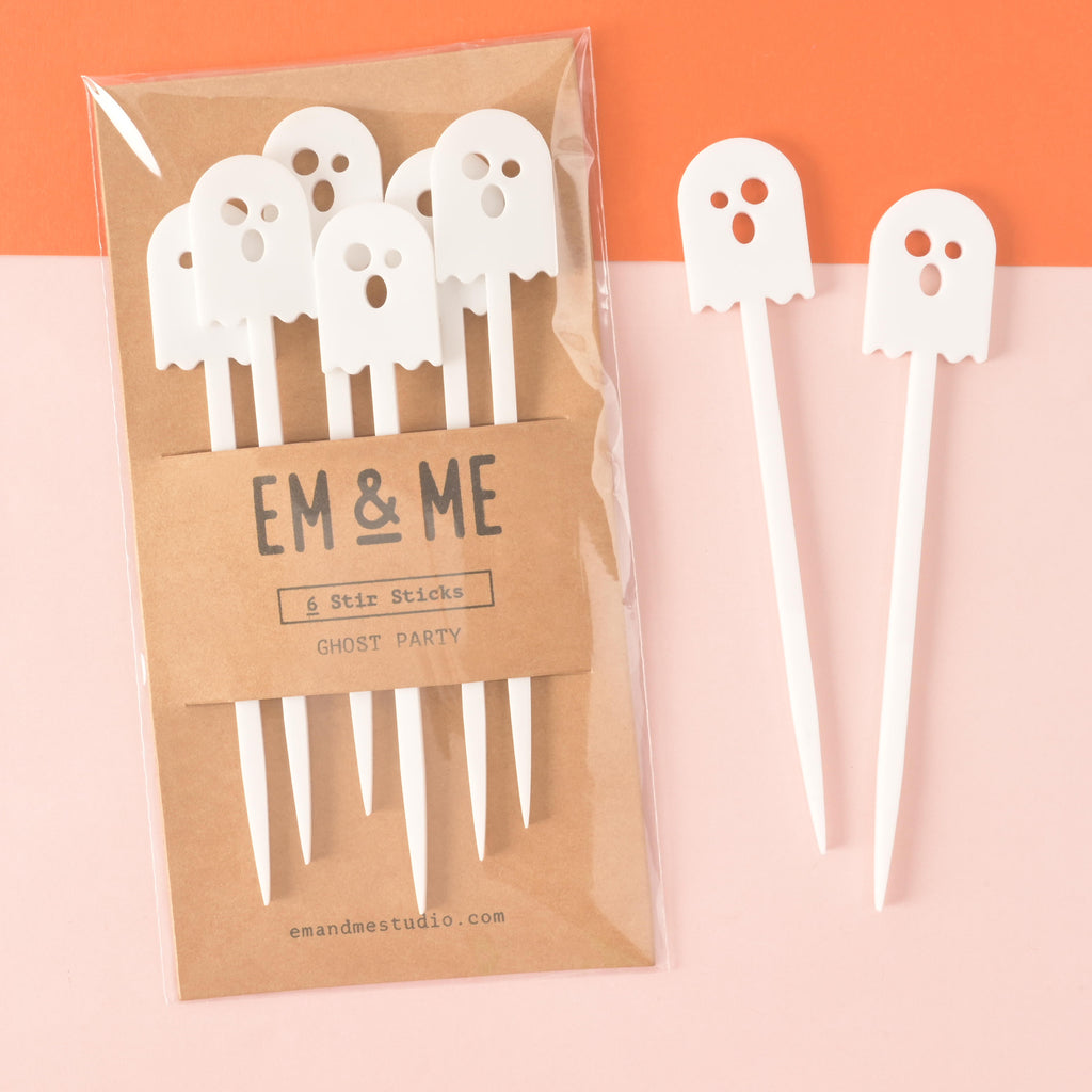 set of six white ghost-shaped cocktail stir sticks packaged on brown craft paper with two more ghosts off to the side on a color blocked background.
