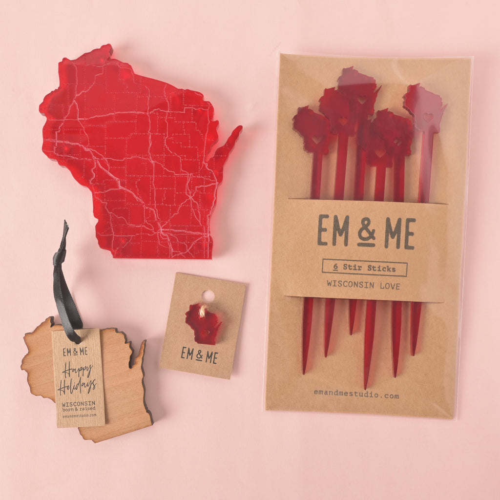 A collection of Wisconsin-themed products, including a large red transparent coaster, a set of six red stirrers in a brown paper package, a wooden "Happy Holidays" ornament, and a small wooden pin. All products are from "EM & ME" and are shown on a solid pink background.