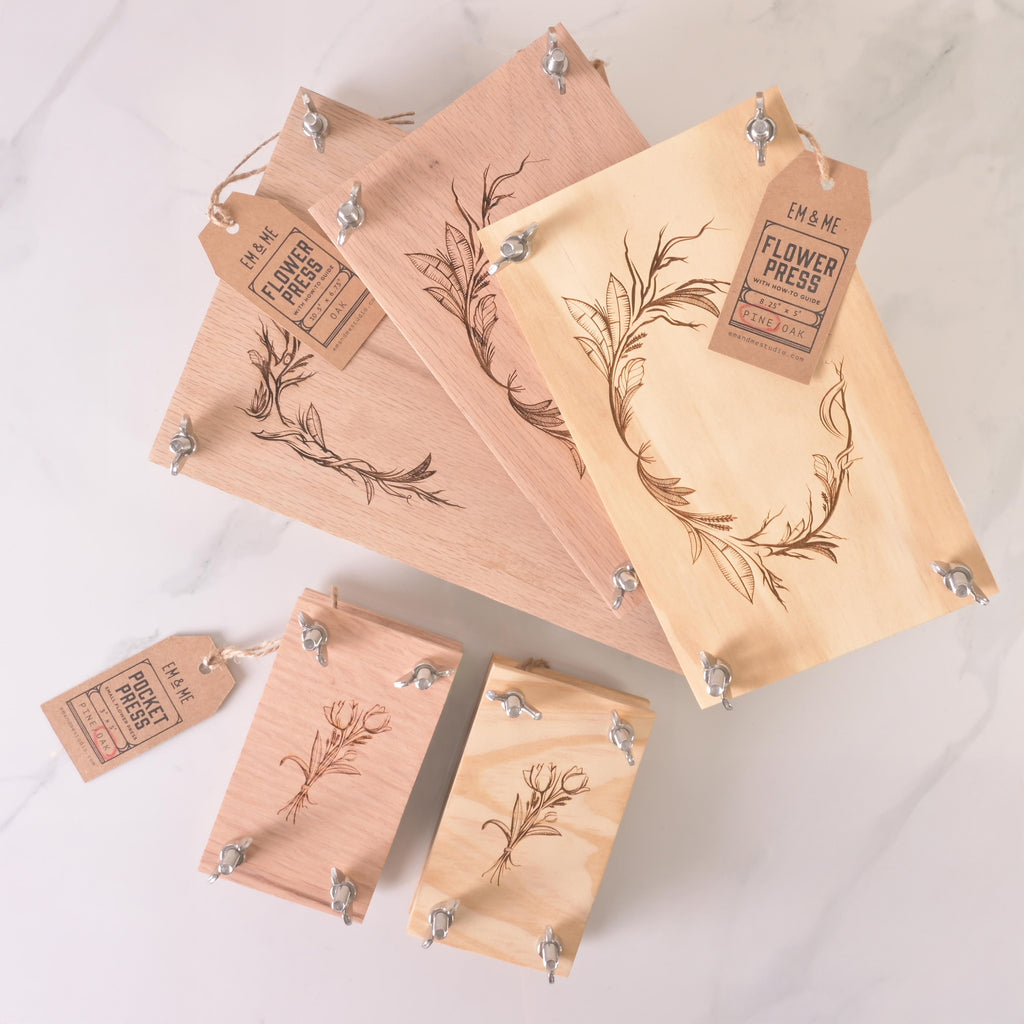 Collection of wooden flower presses in assorted sizes with engraved botanical designs, metal wing nuts, and kraft tags labeled “Flower Press,” displayed on a light background.