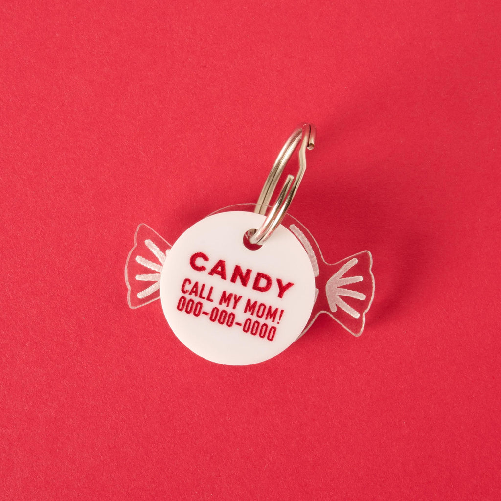A personalized white pet ID tag shaped like a wrapped piece of candy. The tag has the name "CANDY" and a placeholder phone number in red letters. It's attached to a silver ring, perfect for a collar, and is displayed on a solid red background.