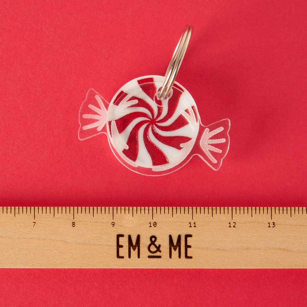 A white and red peppermint candy-shaped charm or keychain in a clear wrapper design. The item is attached to a silver ring. A wooden ruler with the "EM & ME" logo is placed below the charm to show its size. The item is displayed on a solid red background.