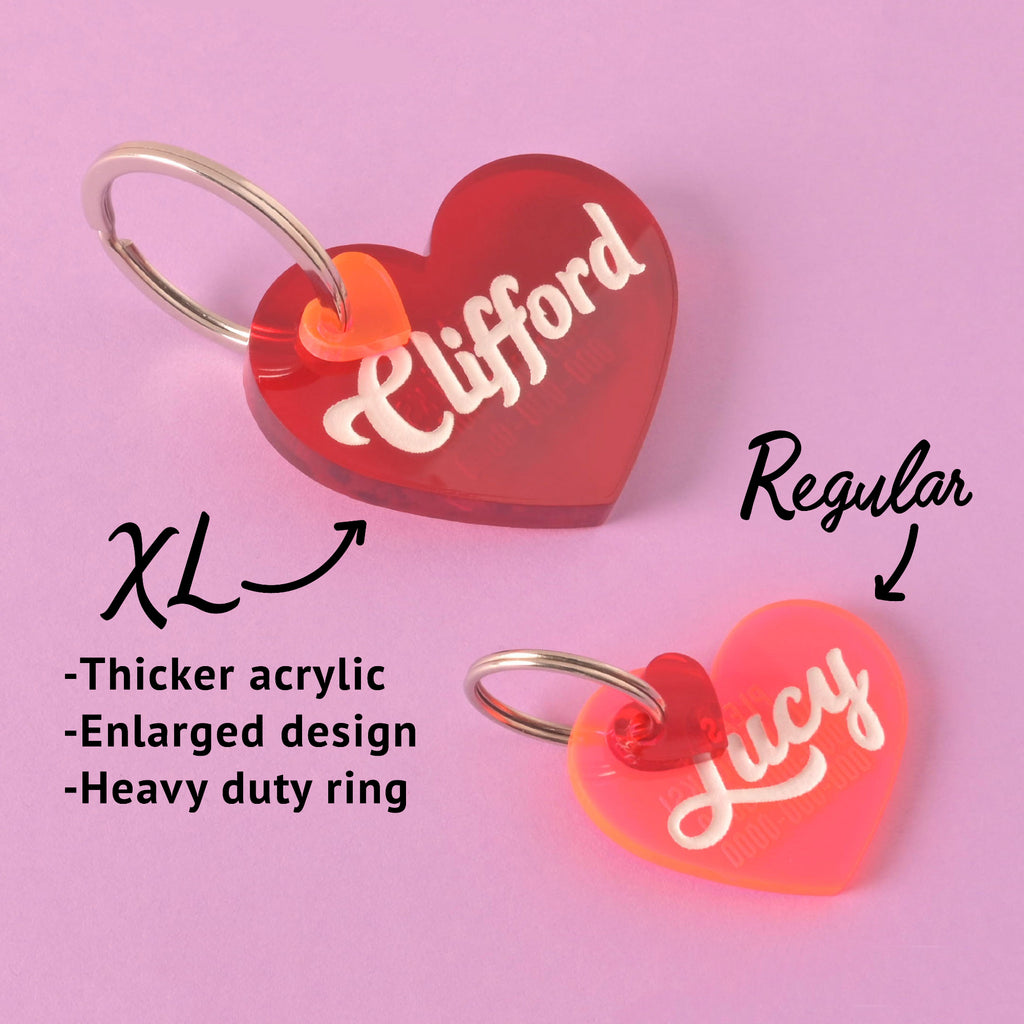 Side-by-side comparison of XL and regular plastic heart-shaped pet ID tags; XL tag for Clifford features thicker acrylic, enlarged design, and a heavy-duty ring, while the regular Lucy tag is shown smaller with standard components, on a purple background