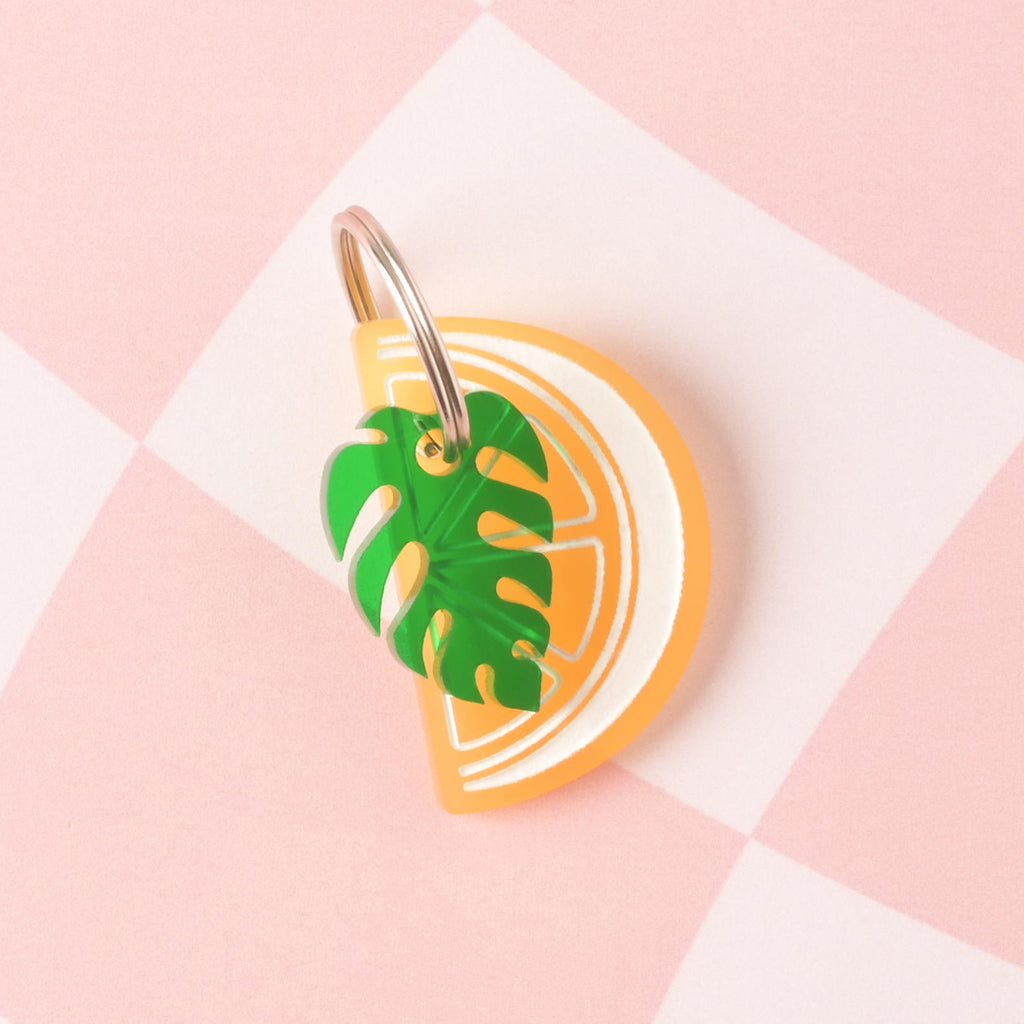 Frosted yellow lemon wedge-shaped personalized pet tag with green monstera leaf mini charm on pink check background.