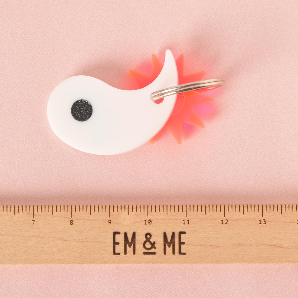 A custom, white, yin yang-shaped pet ID tag with a black circular detail. The tag has a small, spiky, orange starburst charm attached to a shiny silver keyring. A wooden ruler with the logo "EM & ME" is visible at the bottom of the photo, which is set on a solid pink background.