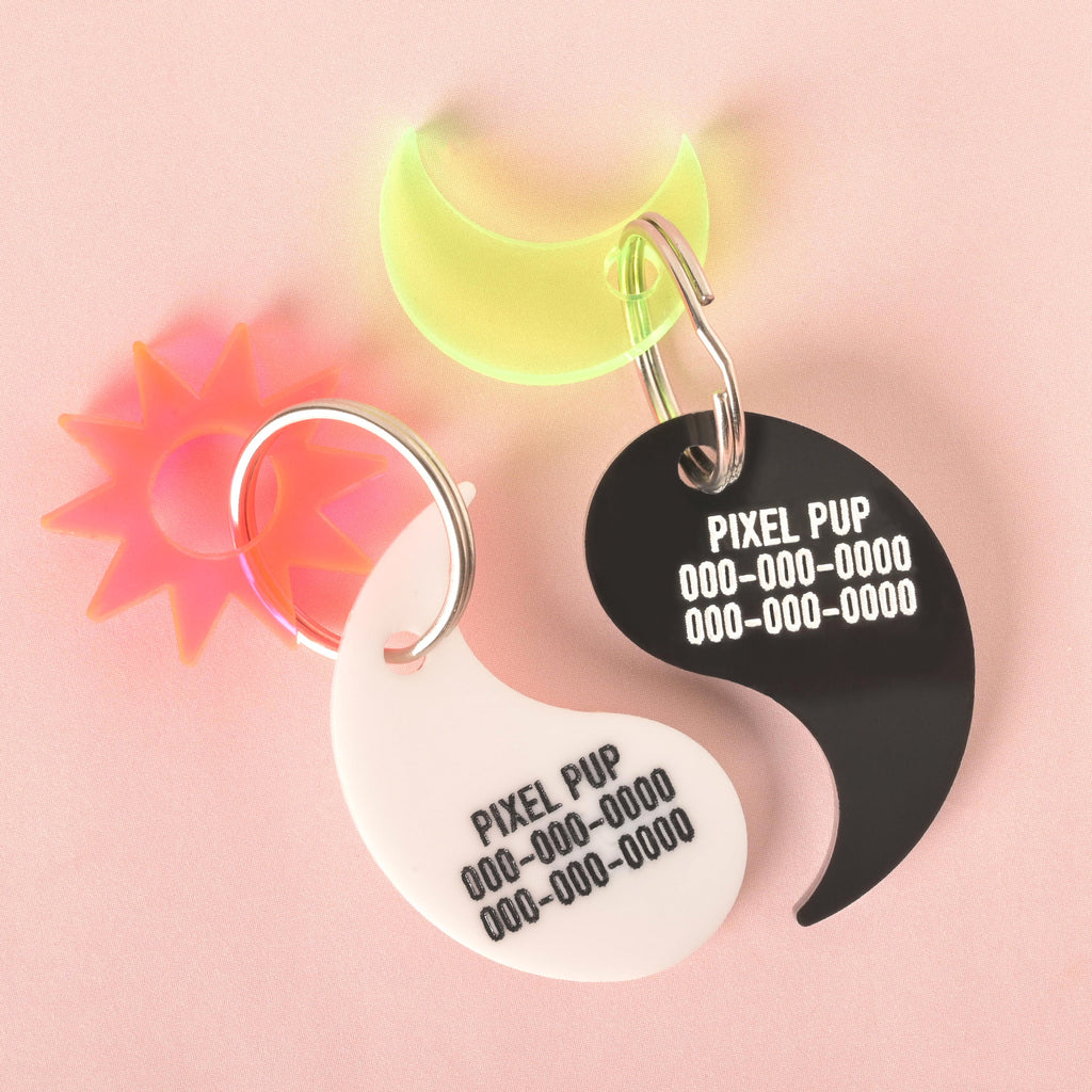 A pair of custom, yin yang-shaped pet ID tags. The white tag and black tag are both engraved with the text "PIXEL PUP" and a phone number. The white tag has a silver keyring with a spiky, orange star charm. The black tag has a silver keyring with a translucent, glow-in-the-dark crescent moon charm. The tags and charms are displayed on a solid pink background.