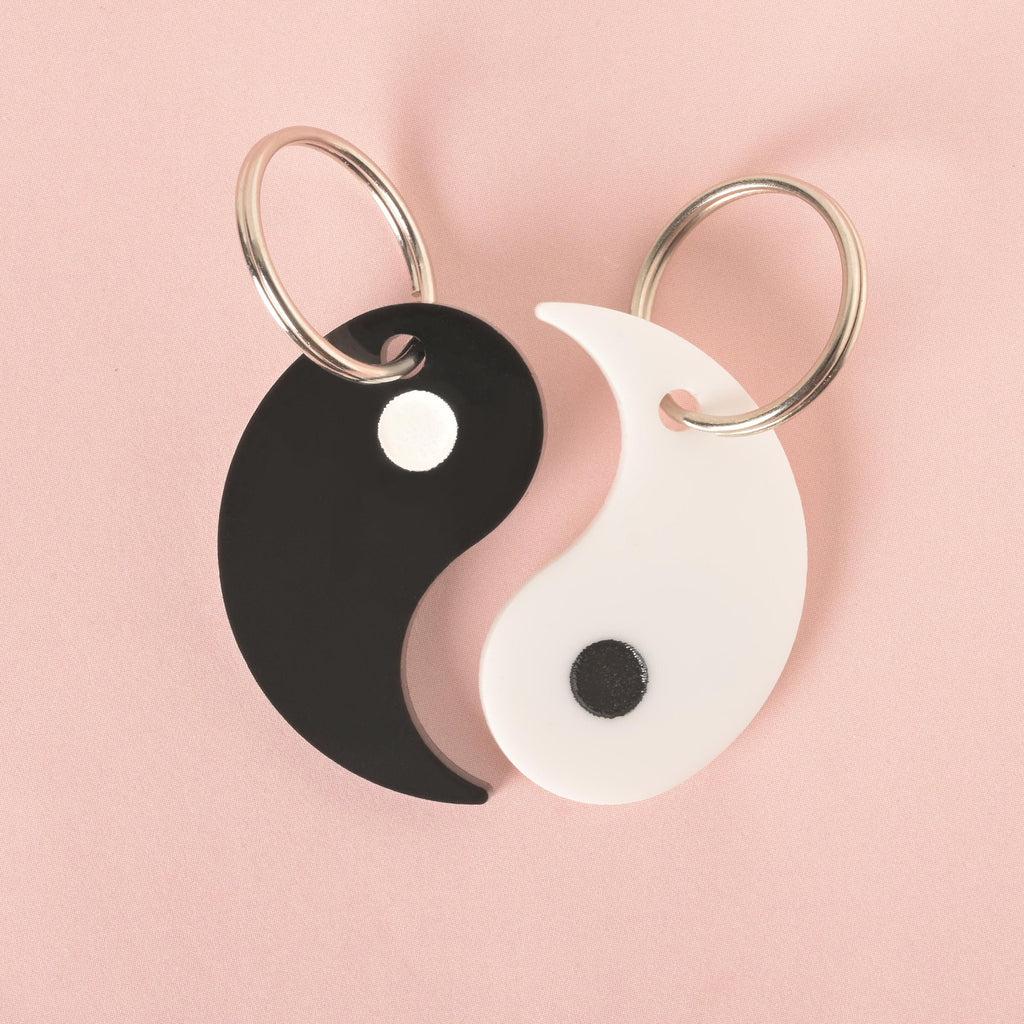 A set of two matching yin yang-shaped pet ID tags. One tag is black with a white circle, and the other is white with a black circle. The tags are connected at the top, each with a silver keyring, and are displayed on a solid pink background.
