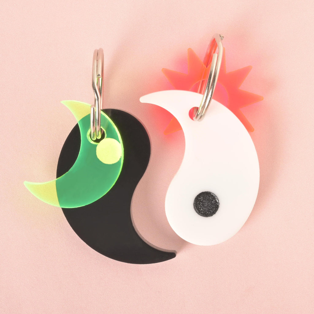 A pair of custom, yin yang-shaped pet ID tags with silver keyrings. The black tag has a translucent, glow-in-the-dark moon charm attached. The white tag has a spiky, orange-red sunburst charm attached. The tags are displayed on a solid pink background.