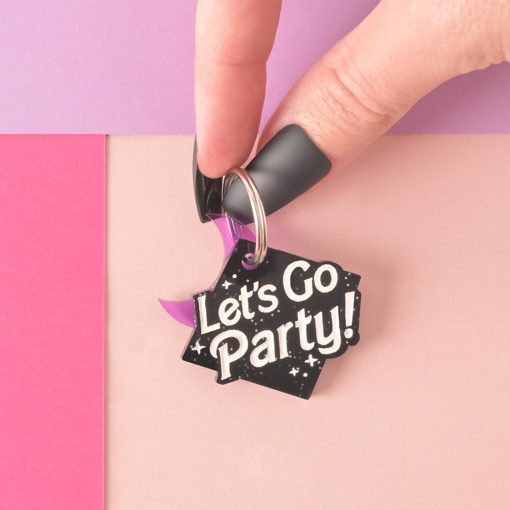 Hand holding a personalized pet tag that reads "let's go party". Glitter black ID tag with moon mini charm held in someones hand with long black fingernails