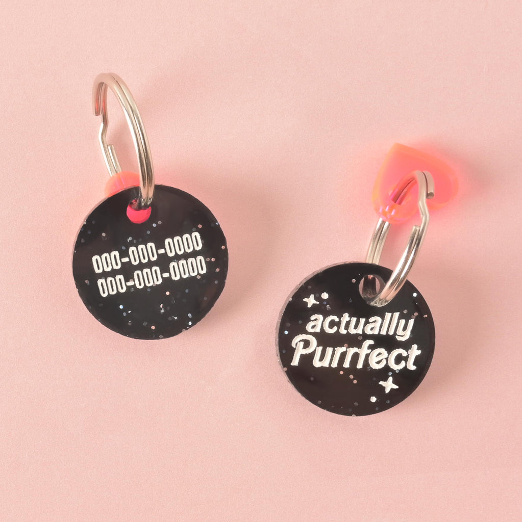 A close-up of a round, black glitter pet tag that reads "actually Purrfect" in white letters. A translucent pink and orange heart charm is attached to the silver ring at the top. The items are displayed on a solid pink background.