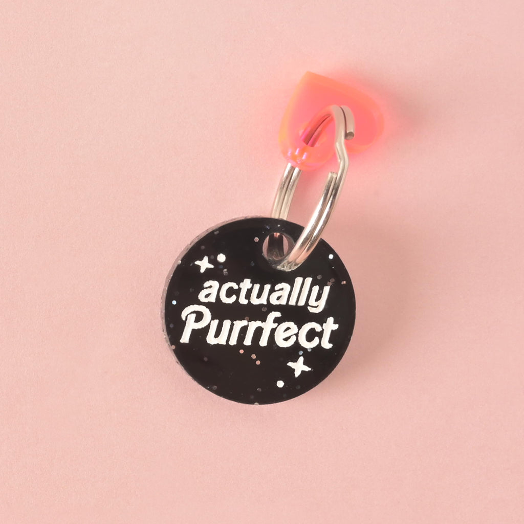 A close-up of a round, black glitter pet tag that reads "actually Purrfect" in white letters. A translucent pink and orange heart charm is attached to the silver ring at the top. The items are displayed on a solid pink background.