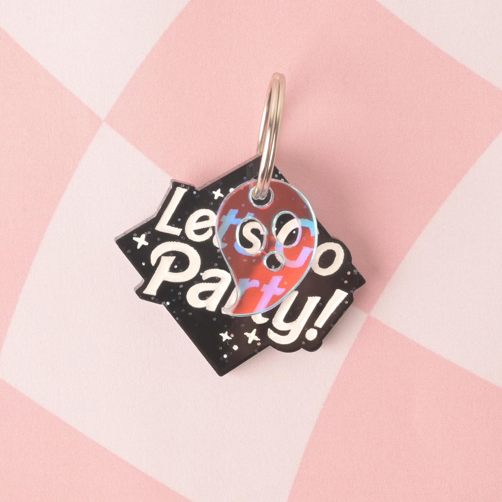 A personalized, black tag in a unique angular shape that says "Let's Go Party!" in white, groovy font. An iridescent ghost charm with a reddish tint is attached to the tag. Both are secured with a silver ring and are resting on a pink and white checkered background.