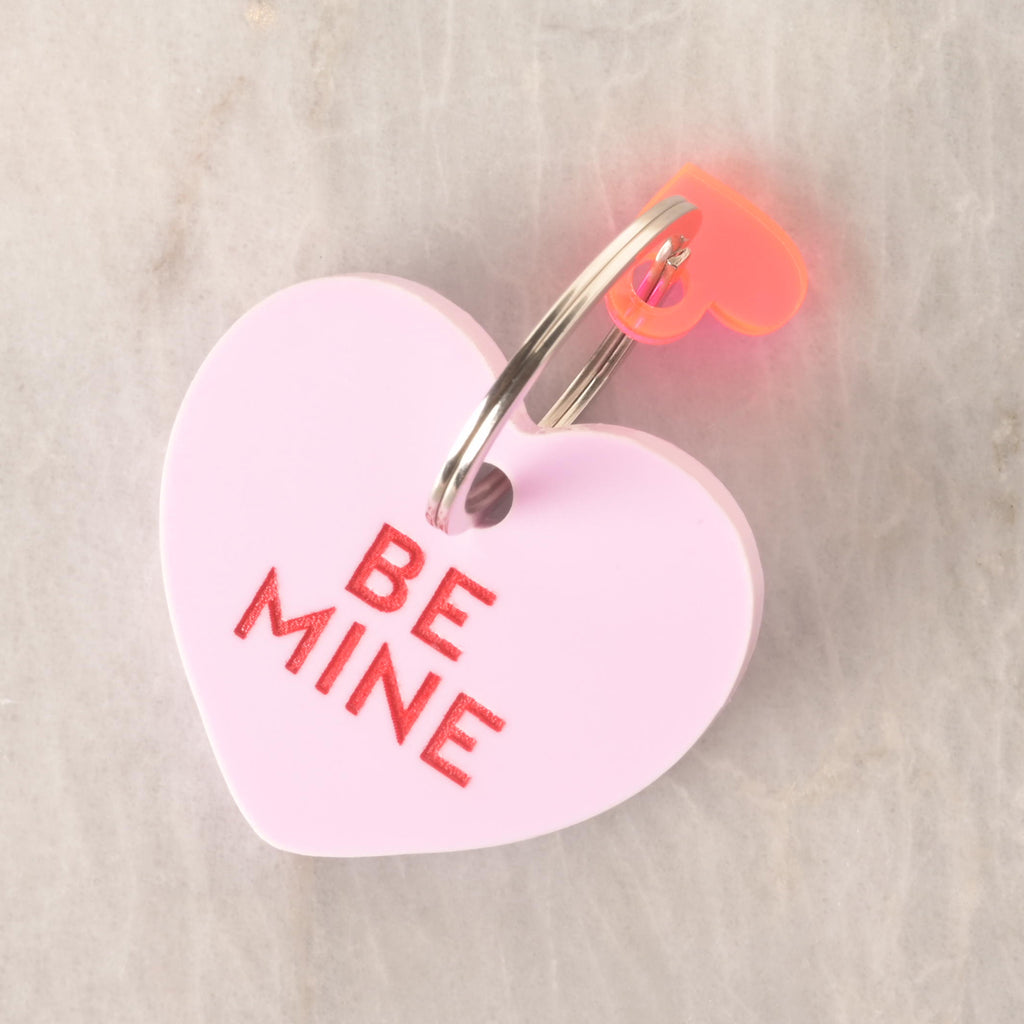 Pink heart-shaped pet ID tag with red engraved text reading ‘Be Mine,’ attached to a red heart keyring, placed on a stone background.