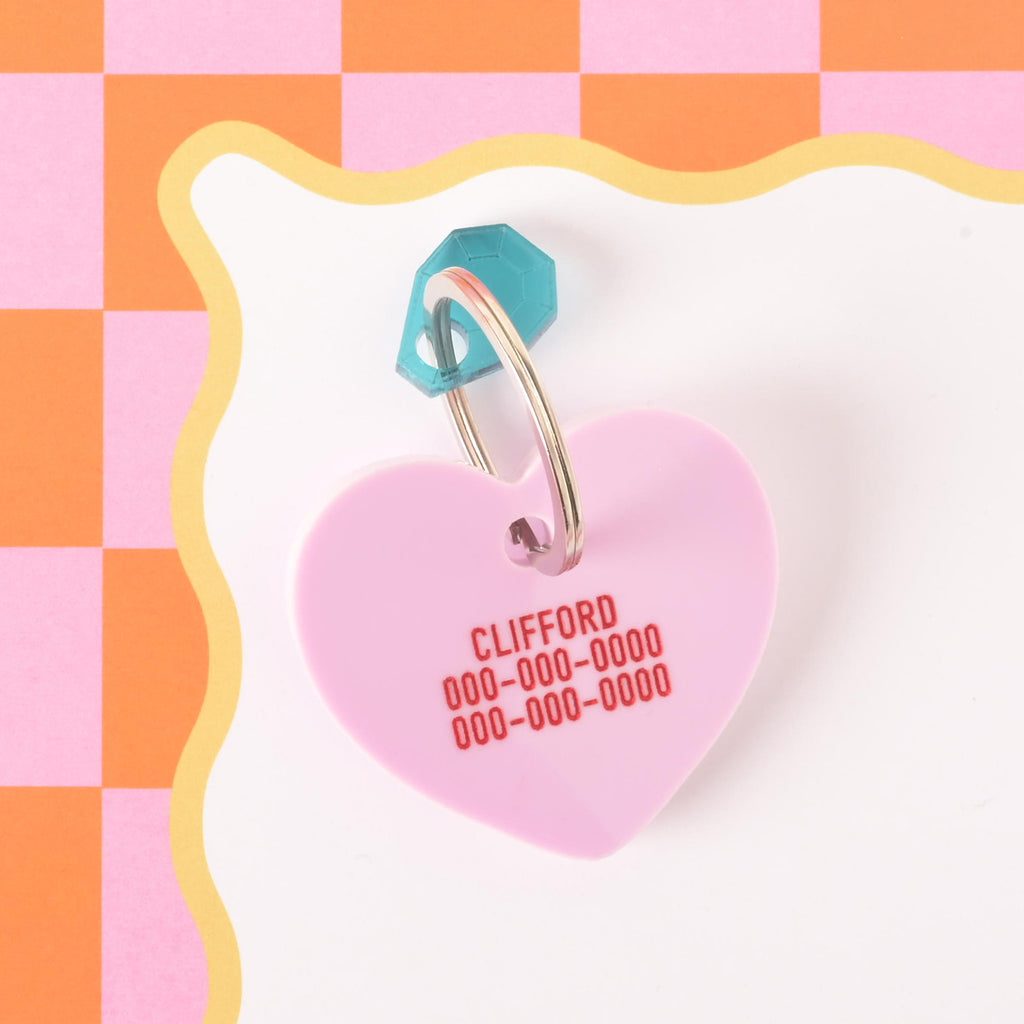 Pink heart-shaped pet ID tag engraved with pet name and phone numbers, attached to a teal gem charm keyring on an orange checkered background.