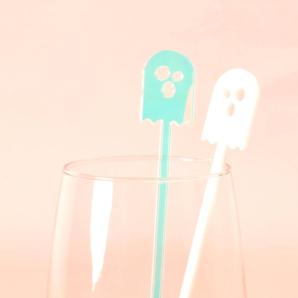 Two ghost-shaped cocktail stir sticks in a clear glass. One stir stick is opaque white the other is iridescent on a solid pink background