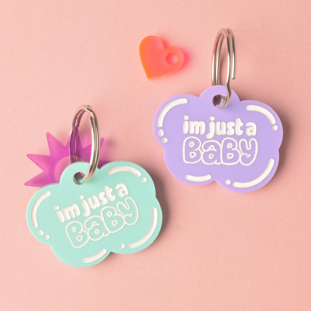 Two acrylic cloud pet ID tags reading “im just a baby”—one mint, one lavender—on silver split rings with heart and star charms on a pink background.