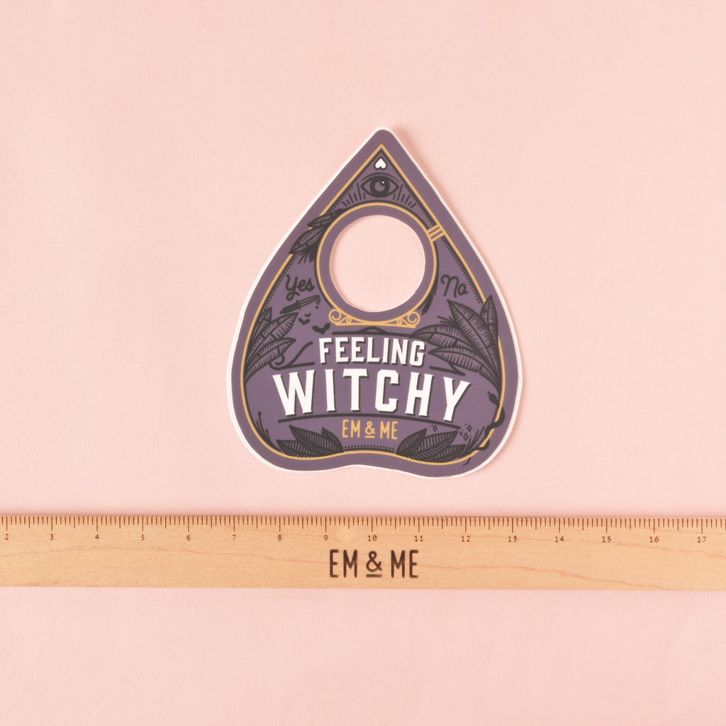 feeling witchy ouija planchet-shaped sticker with wooden ruler that reads "EM & ME" on solid pink background