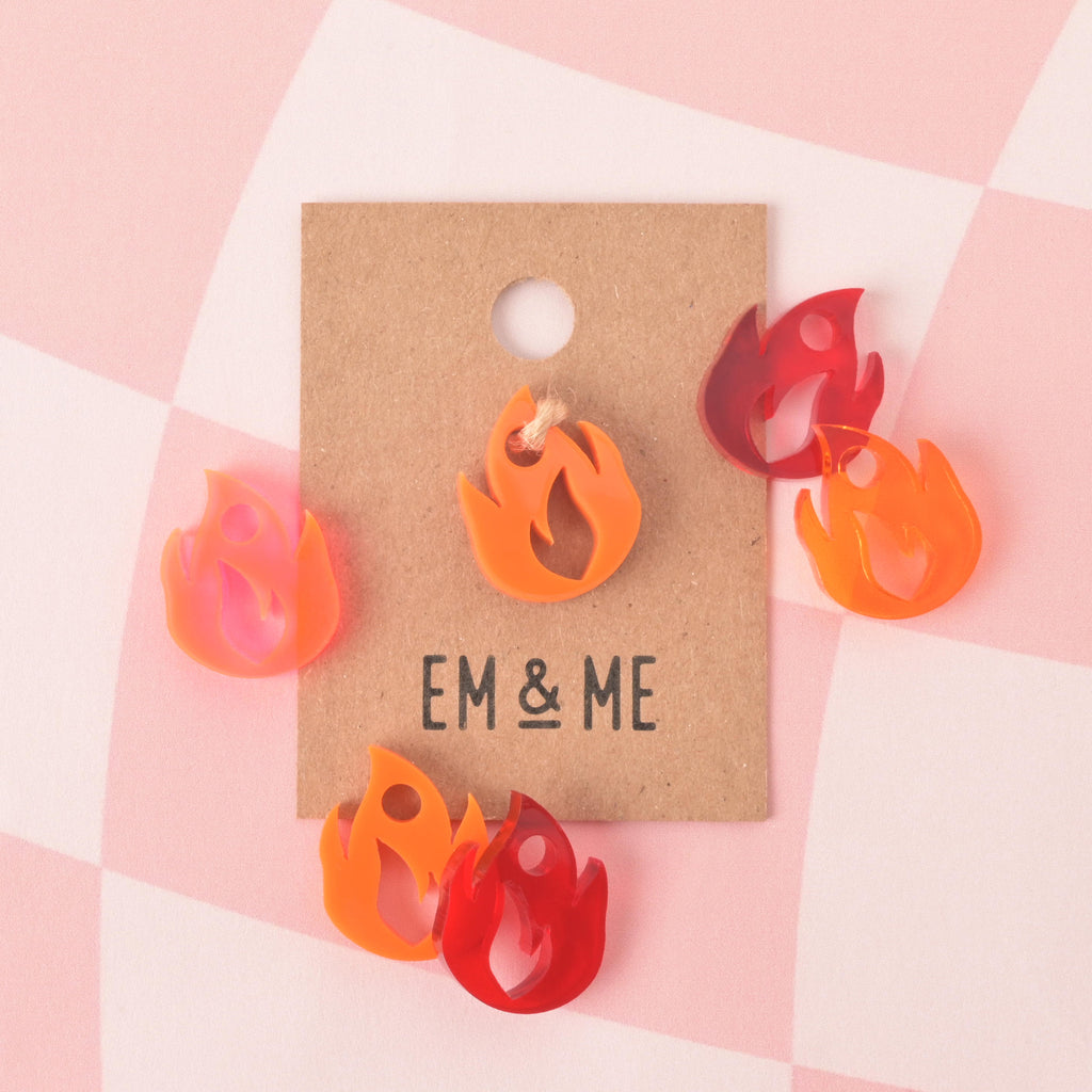 collection of flame-shaped mini charms in red, opaque orange, transparent orange and fluorescent pink colors. one packaged and the others skatered on pink checked background.