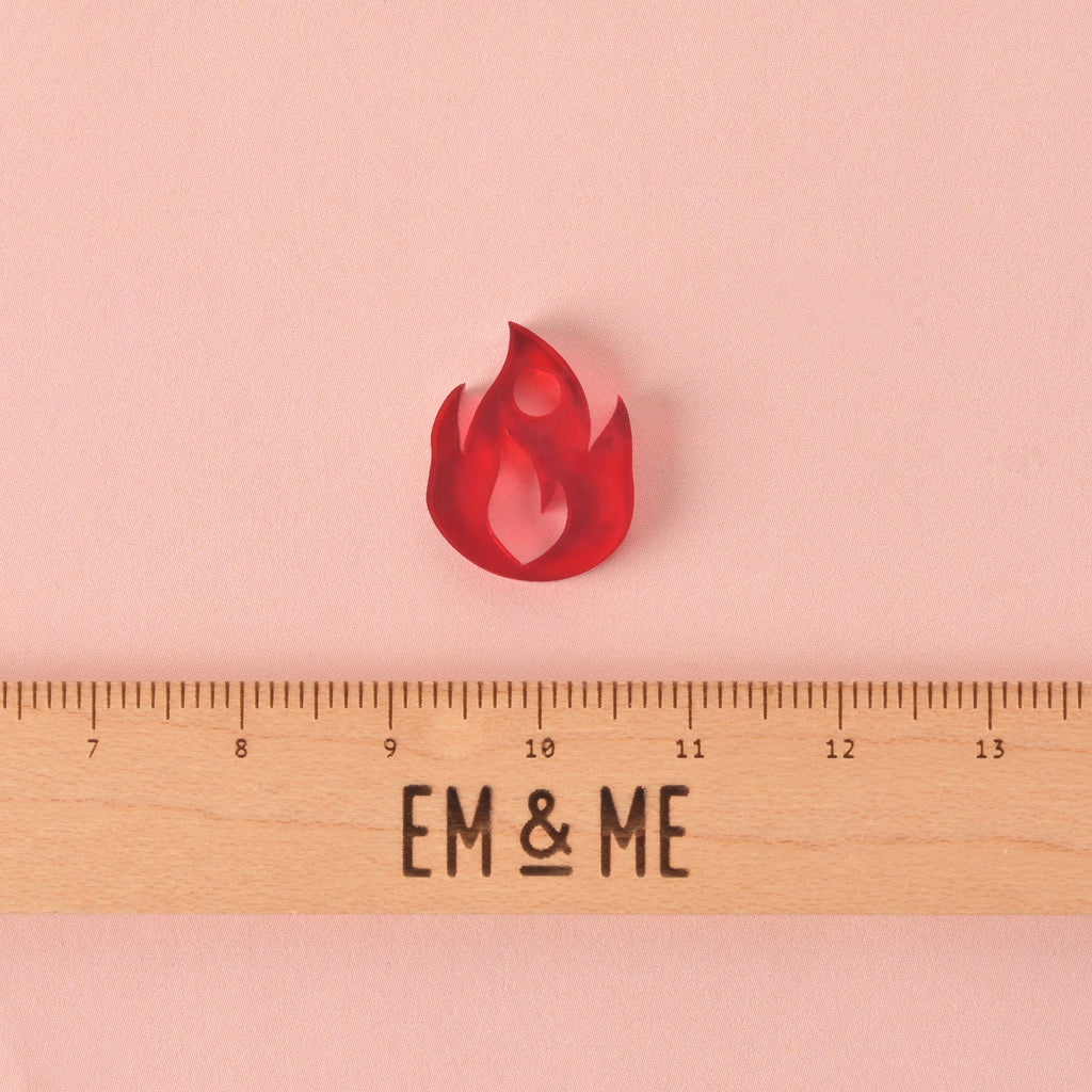 red flame-shaped mini charm with wooden ruler that reads "EM & ME" on a solid pink background.