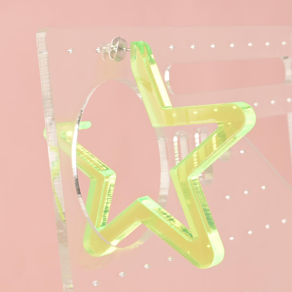 A single large, star-shaped hoop earring made from neon yellow iridescent acrylic. The earring has a silver post and is displayed hanging from a clear earring holder against a solid pink background. The iridescent material reflects a fluorescent green glow.