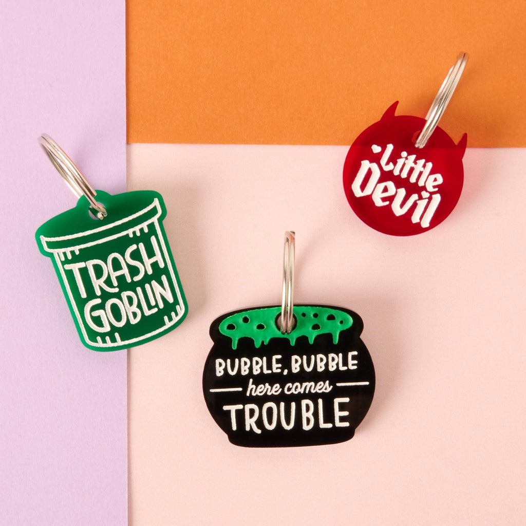 Three funny acrylic pet tags reading ‘Trash Goblin,’ ‘Little Devil,’ and ‘Bubble, Bubble Here Comes Trouble’ displayed on a pastel background