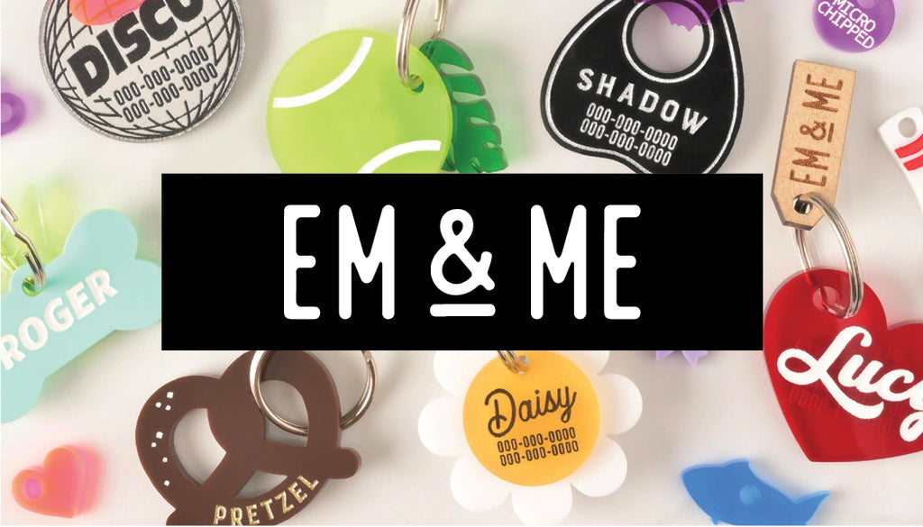 A collection of assorted personalized pet tags in various shapes and colors scattered on a white surface. The tags include a disco ball, a tennis ball, a Ouija planchette, a pretzel, a bone, a flower, and a heart. The items are shown with names like "Roger," "Shadow," and "Daisy." The image has a central black box with the text "EM & ME."