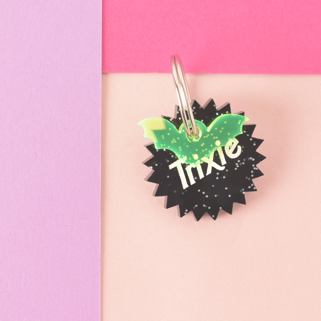 Glitter black barbie inspired sunburst-shaped personalized pet tag that reads "Trixie" with fluorescent green bat mini charm on color blocked background.