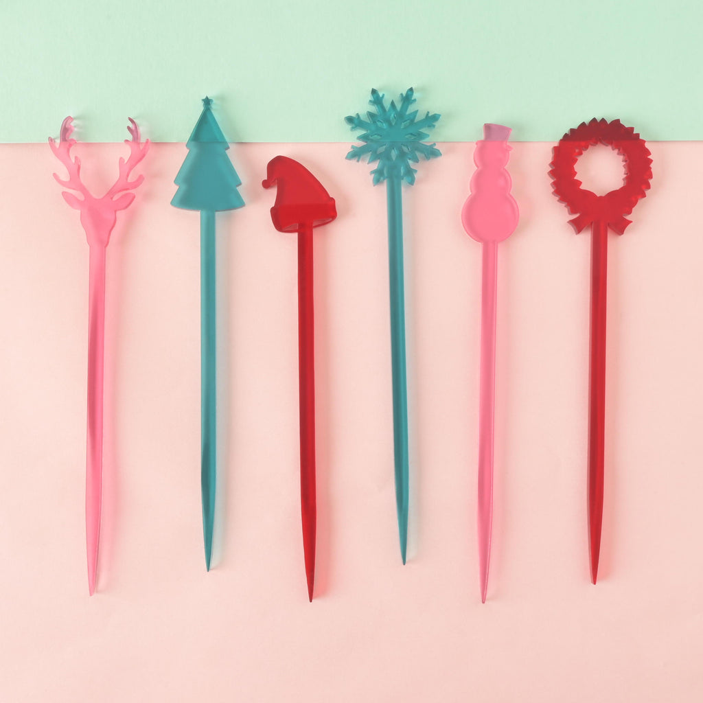 A set of six colorful Christmas-themed cocktail stirrers, arranged side-by-side on a light pink and mint green background. The stirrers feature various holiday shapes: a pink reindeer, a teal Christmas tree, a red Santa hat, a teal snowflake, a pink snowman, and a red wreath.