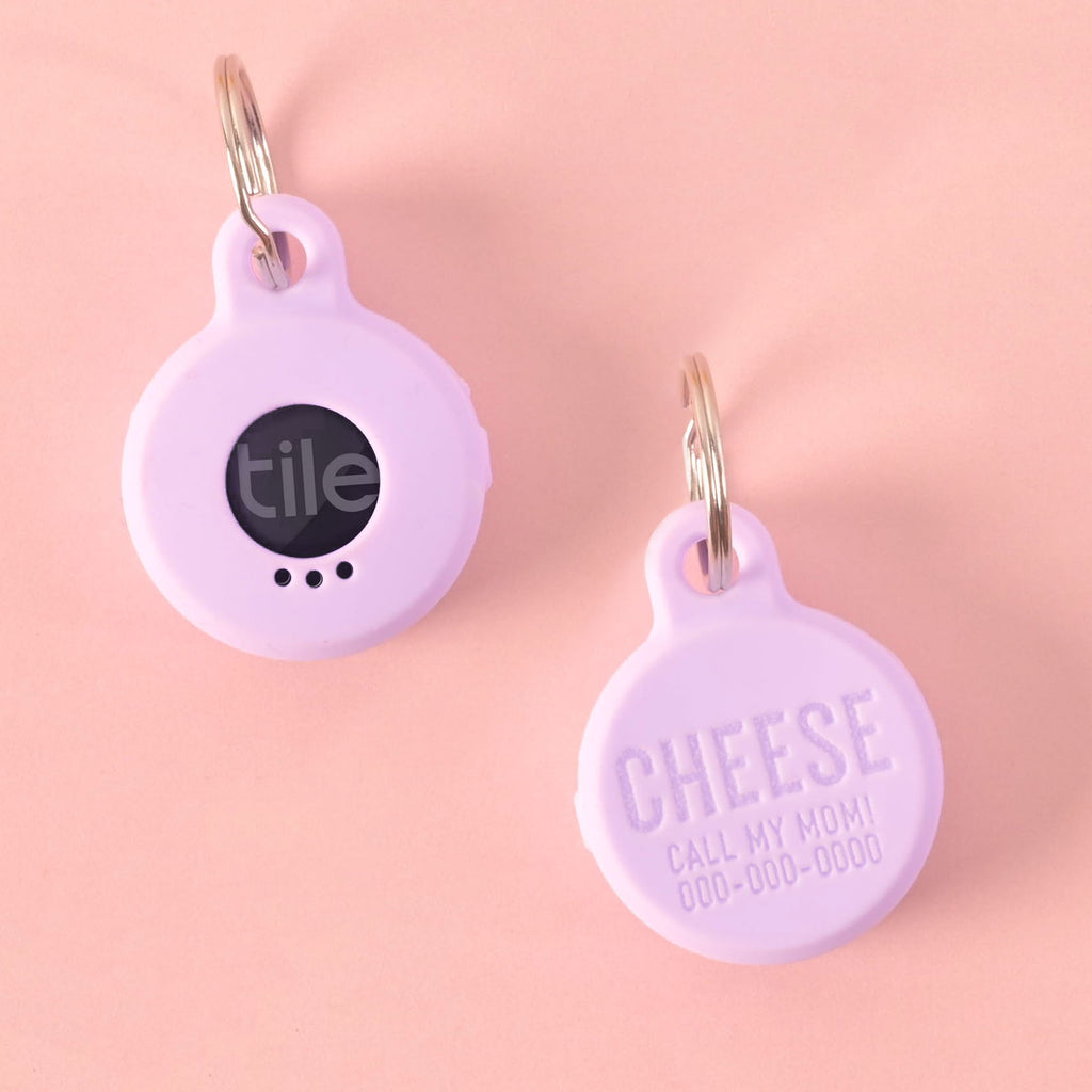 A pair of purple silicone pet tags designed to hold a Tile tracker. The front of one tag shows the Tile logo, while the other is personalized with the name "CHEESE" and a placeholder phone number. Both tags are attached to a silver ring for a collar and are displayed on a solid pink background.