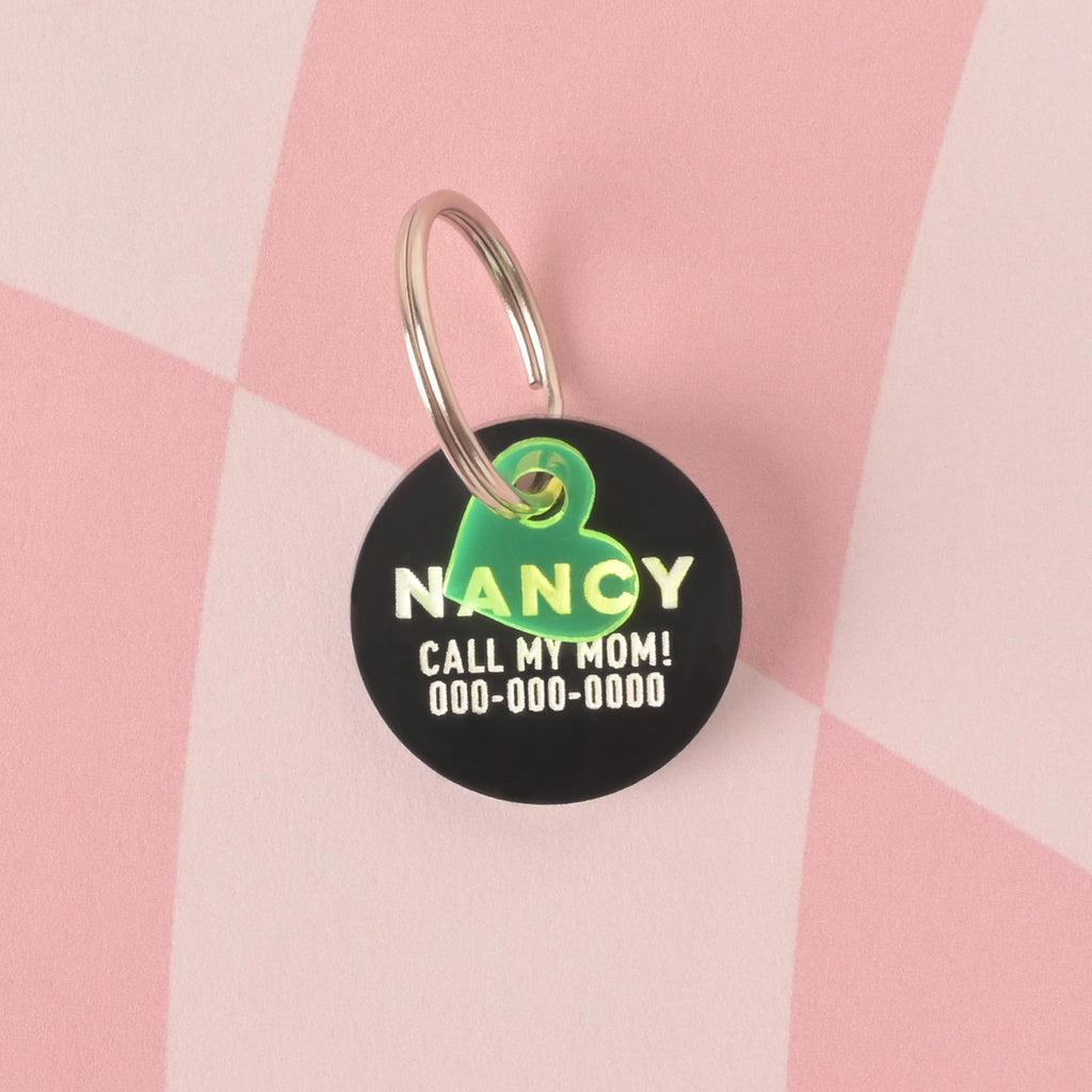 A personalized opaque black round pet tag with the name "NANCY" and a placeholder phone number in white letters. A small, translucent neon green heart charm is attached to the tag's silver ring. The items are displayed on a pink and white checkered background.