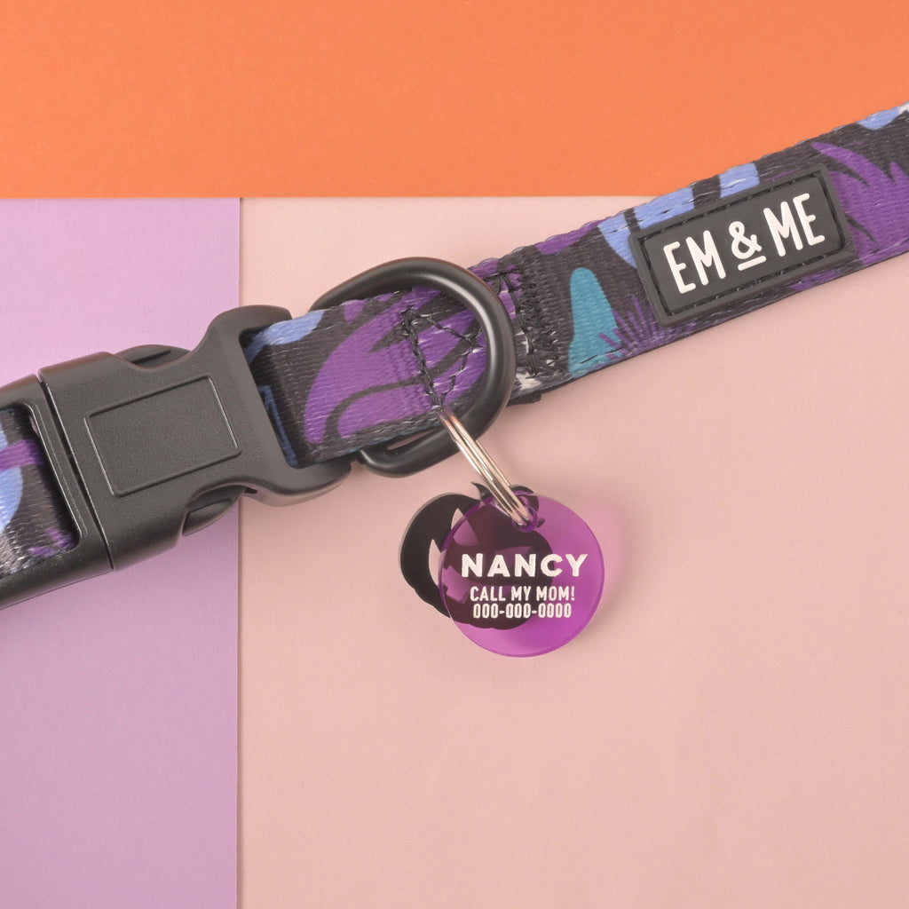 A personalized purple round pet tag with the name "NANCY" and a placeholder phone number attached to a black and purple patterned dog collar. The tag has a shiny, mirrored finish and is secured with a silver ring. The items are displayed on a background of orange, light purple, and pink panels.