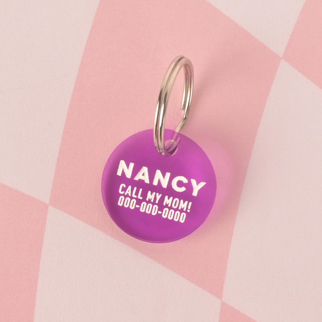 A personalized, purple round pet tag with the name "NANCY" and the phrase "CALL MY MOM!" with a placeholder phone number on it. The tag is attached to a silver ring, perfect for a collar, and is displayed on a pink and white checkered background.