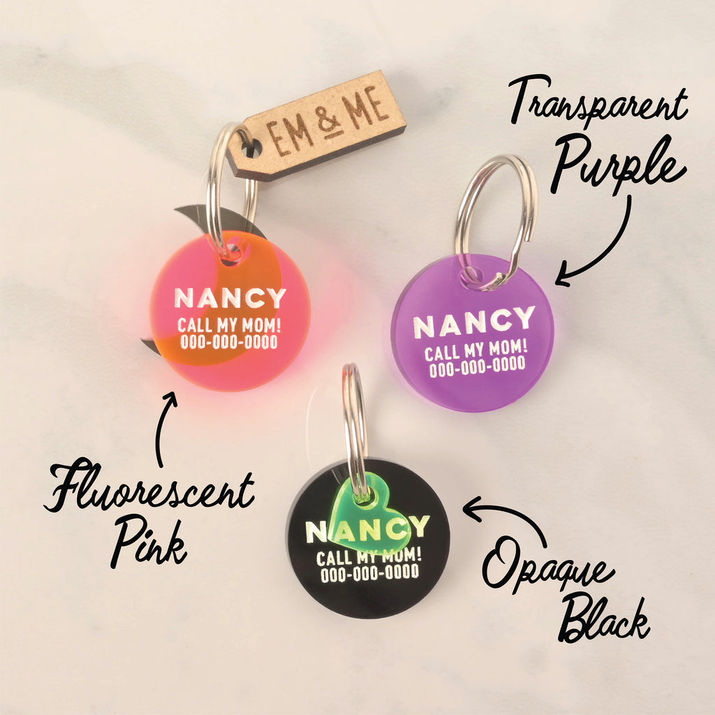 A set of three personalized round pet tags for "NANCY," shown in different colors and finishes. The tags include a Fluorescent Pink one, a Transparent Purple one, and an Opaque Black one. All tags have a placeholder phone number and are attached to a silver ring for a collar. A small wooden "EM & ME" tag and a translucent green heart charm are also visible, highlighting the color options.