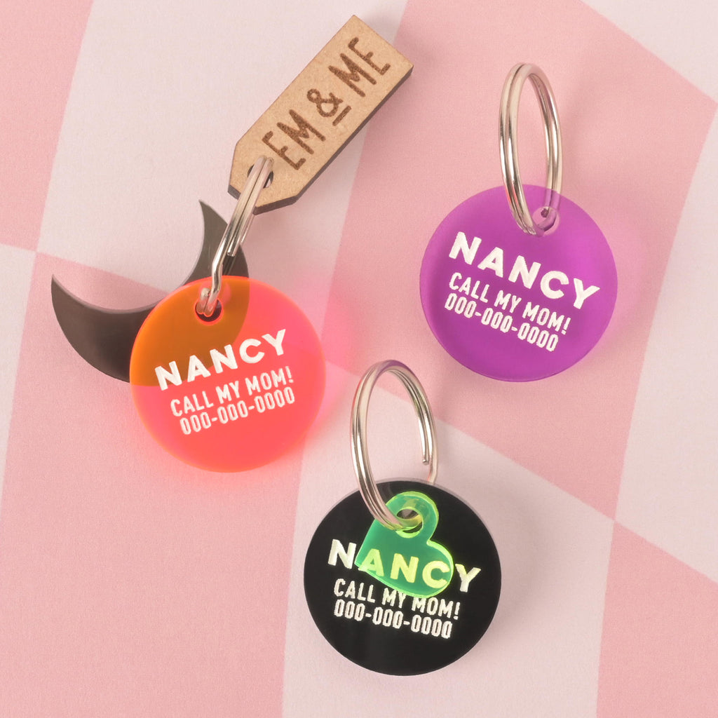 A set of three personalized round pet tags in orange, purple, and black. All tags feature the name "NANCY" and a placeholder phone number, with the phrase "CALL MY MOM!". The black tag includes a small, translucent green heart charm. A wooden "EM & ME" tag is also attached to the orange tag. The items are displayed on a pink and white checkered background.
