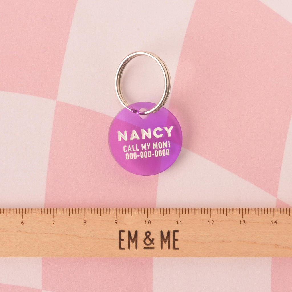 A personalized transparent purple round pet tag with the name "NANCY" and a placeholder phone number on it. The tag is attached to a silver ring, perfect for a collar. A wooden ruler with the "EM & ME" logo is placed below the tag to show its size. The items are displayed on a pink and white checkered background.