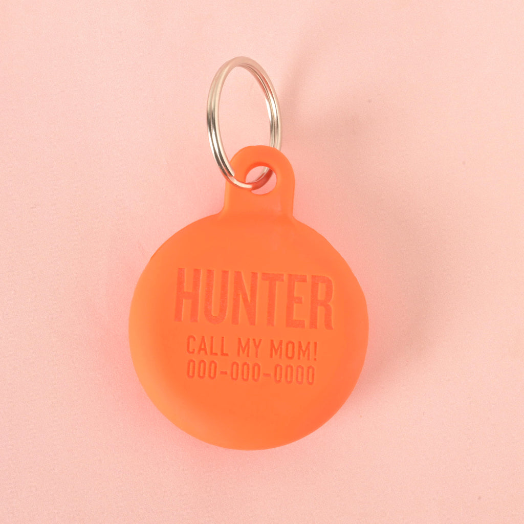 A close-up shot of a custom, round, orange pet ID tag. The tag is engraved with the name "HUNTER," the phrase "CALL MY MOM!", and a phone number below it. It's attached to a shiny silver keyring and is displayed on a solid pink background.