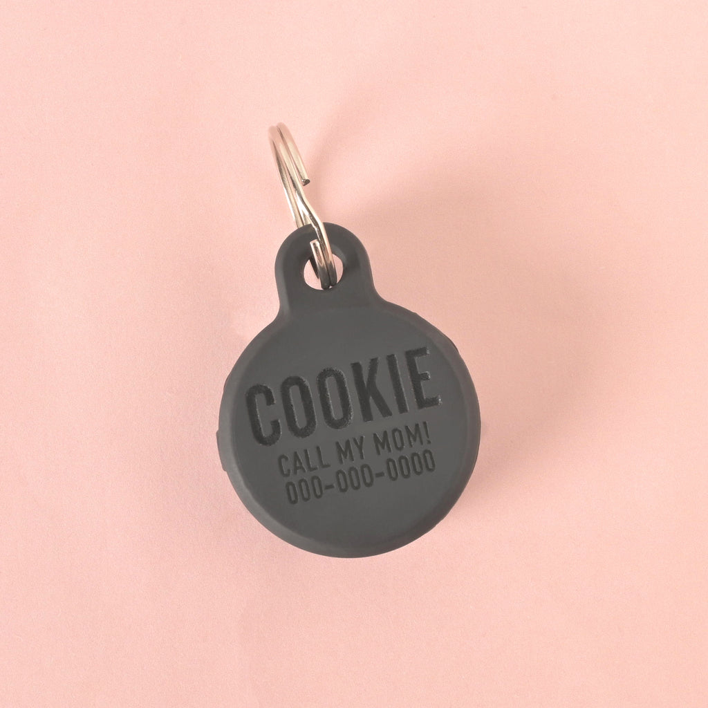 A close-up shot of a custom, round, Black pet ID tag. The tag is engraved with the name "COOKIE," the phrase "CALL MY MOM!", and a phone number. It is attached to a shiny silver keyring and is displayed on a solid pink background.