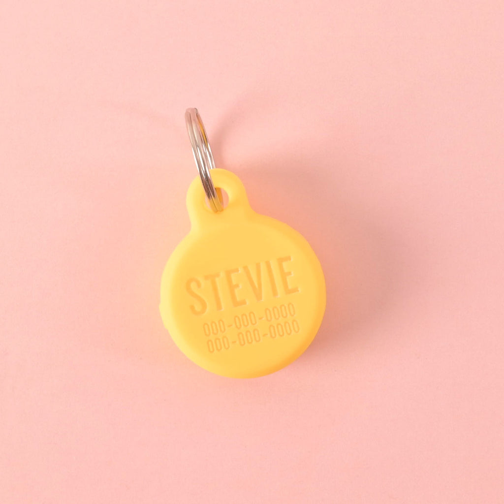 A personalized yellow Tile Sticker holder for pets that says "STEVIE" and has a placeholder phone number on it. The holder is attached to a silver ring, perfect for a collar, and is displayed on a solid pink background.