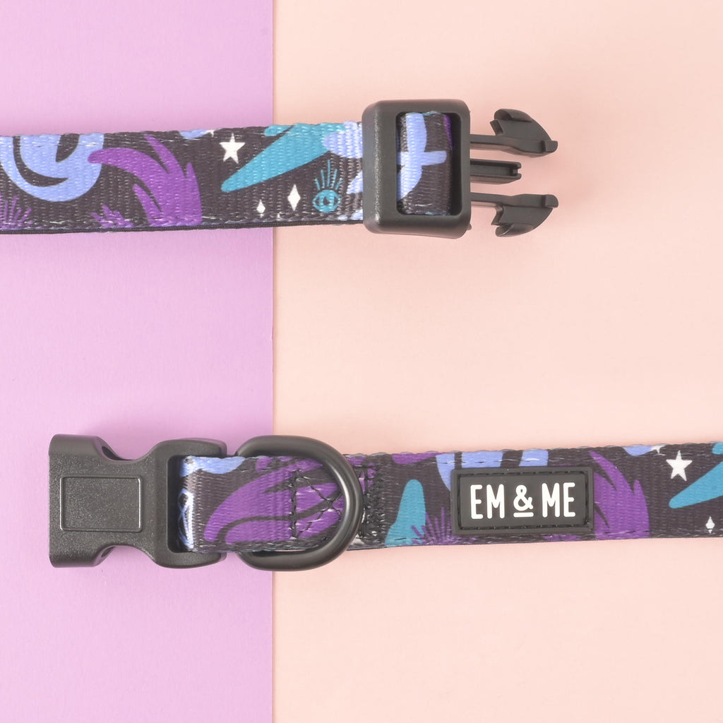 Close-up of adjustable dog collar in purple and teal celestial print showing black side-release buckle open and sturdy D-ring; EM & ME logo; pink and lavender background.