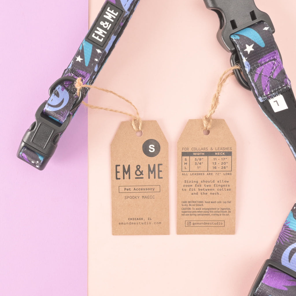 Close-up flat lay of EM & ME “Spooky Magic” adjustable dog collar in purple and teal celestial print with two kraft hang tags; one shows brand and size S, the other shows collar size chart: S 11–17 in, M 13–20 in, L 16–26 in; pink and lavender background.