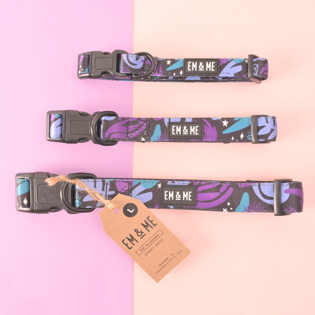 Flat lay of three adjustable dog collars in a purple-teal celestial pattern with stars; sizes S, M, and L with side-release buckles and D-rings; EM & ME label; pink background with kraft size tag.