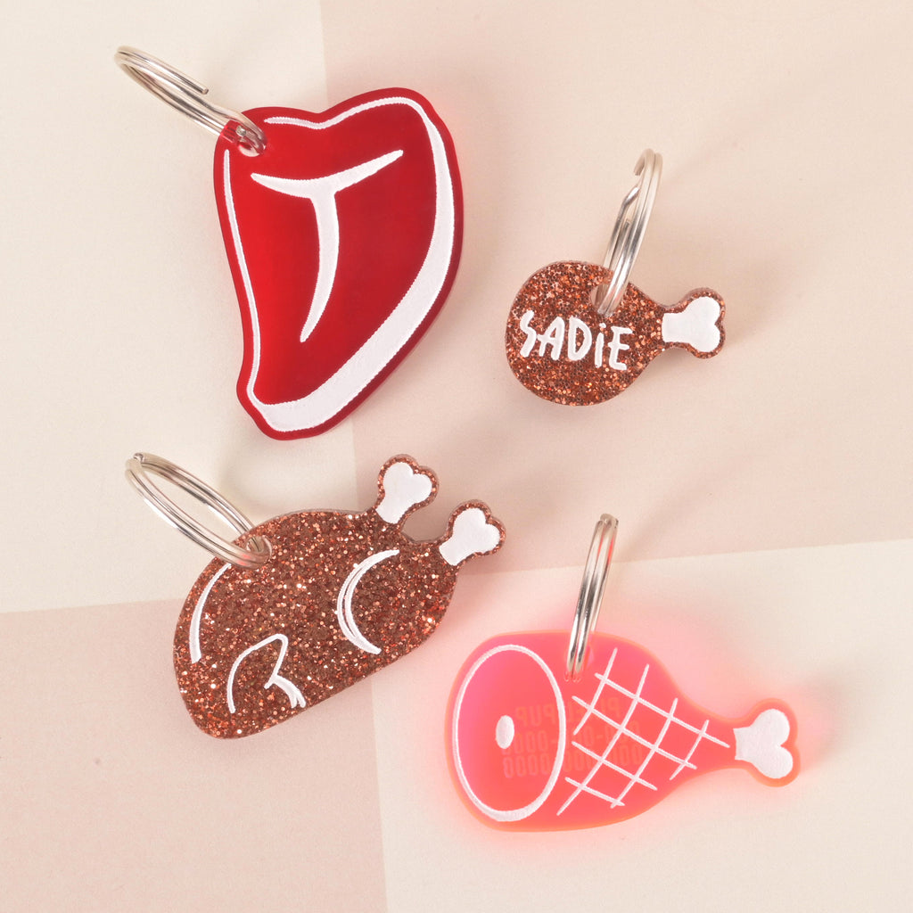Set of meat-themed pet ID tags including a red steak, glittery drumstick, glitter bone-shaped tag with “Sadie,” and a pink ham tag, customizable name charms for cats and dogs.