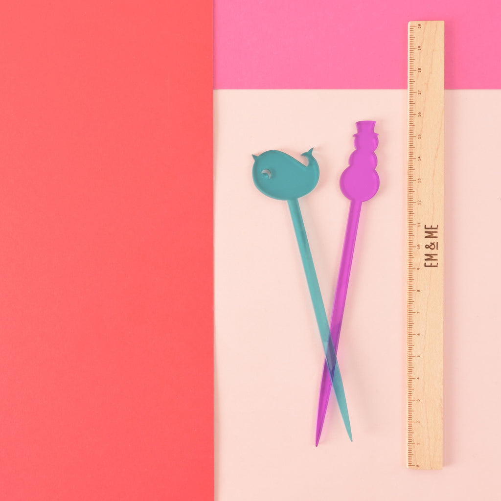 teal narwhal-shaped and purple snowman-shaped stir sticks next to a wooden ruler on color blocked background