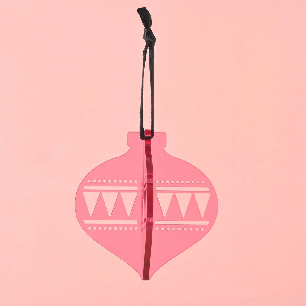 A single 3D interlocking acrylic Christmas ornament in a bright pink color, featuring white engraved geometric and triangle patterns. The ornament hangs from a black ribbon against a solid pink background. Acrylic Christmas ornaments, bright colors and non-breakable